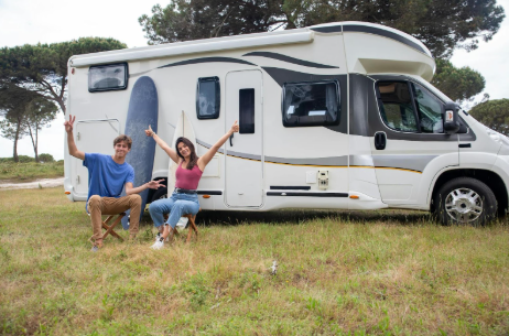 Couple sitting in the grass outside motorhome with a surfboard leaned against the vehicle.